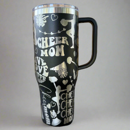 A 40 oz stainless steel tumbler with a 'Cheer Mom' theme, featuring laser engraved text and symbols on a black background, displayed on a surface with a blurred background.